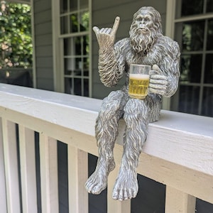 May include: A silver-colored Bigfoot statue with a beer mug, gesturing a hand sign. The statue is seated on a white railing, with a house and windows in the background. The beer mug is filled with a light-colored liquid.