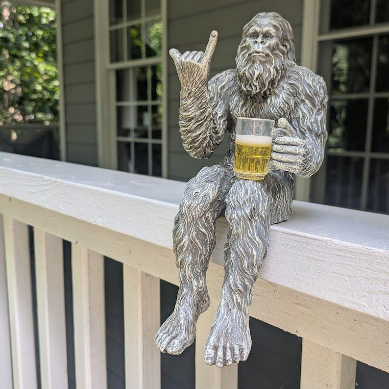 May include: A silver-colored Bigfoot statue with a beer mug, gesturing a hand sign. The statue is seated on a white railing, with a house and windows in the background. The beer mug is filled with a light-colored liquid.