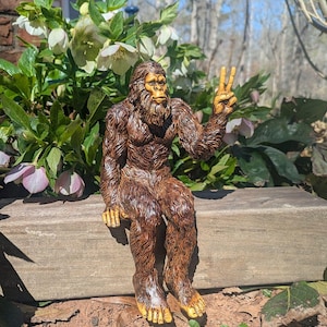 May include: A brown, furry Bigfoot figurine sitting on a wooden ledge. The creature has a textured coat, a detailed face with a beard, and is holding its left hand up in a peace sign gesture. The figure is depicted with visible toes and fingers.