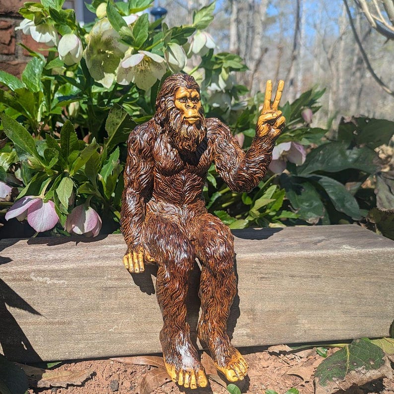 May include: A brown, furry Bigfoot figurine sitting on a wooden ledge. The creature has a textured coat, a detailed face with a beard, and is holding its left hand up in a peace sign gesture. The figure is depicted with visible toes and fingers.