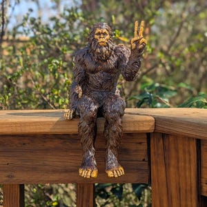 May include: A brown, furry, ape-like creature figurine with a textured coat and light-colored hands and feet. The figure is depicted in a seated position with one arm raised, displaying a hand gesture with two fingers extended in a V-shape.