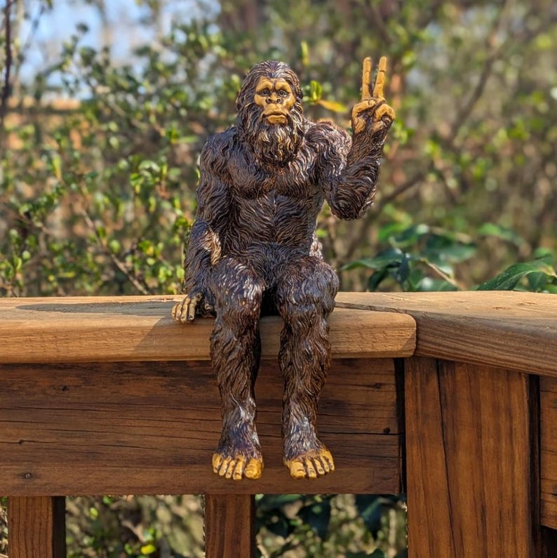 May include: A brown, furry, ape-like creature figurine with a textured coat and light-colored hands and feet. The figure is depicted in a seated position with one arm raised, displaying a hand gesture with two fingers extended in a V-shape.