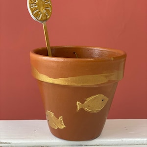 Puede incluir: Una maceta marrón con detalles dorados y diseños de peces. Un marcador de plantas dorado con la palabra "GIULIETTA" en su interior. La maceta está sobre una superficie blanca contra un fondo rojo.