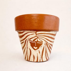 Peut inclure: Un pot de fleur en céramique de couleur terre cuite avec un motif peint à la main. Le motif représente un visage blanc aux yeux fermés et aux cheveux flottants. Le pot a un large bord et un corps conique.