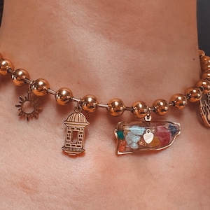 Op de afbeelding: Gouden kralen choker ketting met meerdere bedels, waaronder een klein huis, een zon en een heldere bedel gevuld met gekleurde stenen.