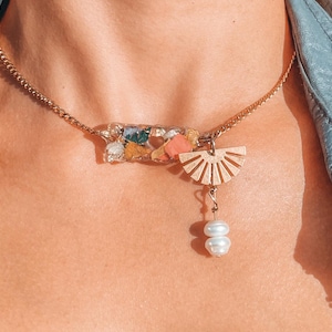 May include: A gold chain necklace with a resin pendant filled with colorful pieces of dried flowers and a gold sun charm with two white pearl beads hanging from it.