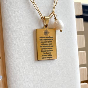 May include: A gold-colored necklace with a rectangular pendant and a pearl charm. The pendant has engraved text and a floral design. The chain is a link style.
