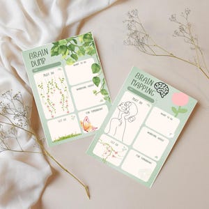 May include: Two rectangular paper planners with light green borders. The left planner is titled Brain Dump and features hanging vines, a butterfly, and grass. The right planner is titled Brain Mapping with a brain icon, a line-art face, and a pink flower.