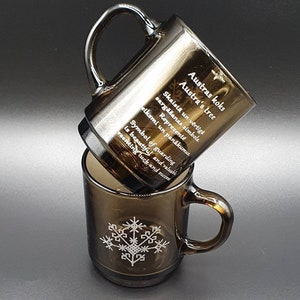 May include: Two brown glass mugs stacked on top of each other. The top mug has text that reads "Austra's tree" and "Symbol of guarding." The bottom mug has a white snowflake design. The mugs have a handle.