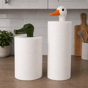 Puede incluir: Dos rollos de papel de cocina blancos con remates decorativos. Un rollo tiene una cabeza de cocodrilo verde y el otro una cabeza de pato blanca con pico naranja. Ambos rollos presentan un patrón de diamantes texturizado en el papel.