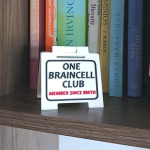 May include: A shelf displays a collection of books with various colored spines. A white sign with black text that reads "ONE BRAINCELL CLUB MEMBER SINCE BIRTH" is in front of the books. A Jenga game is stacked on the right side of the shelf.