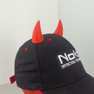 May include: Black baseball cap with red trim and red devil horns. The cap has a white logo that says "Nokta Detection Technology".