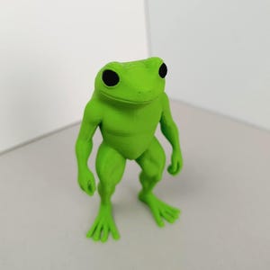 Puede incluir: Una figurita de rana verde con ojos negros. La rana está de pie con los brazos y las piernas extendidos.