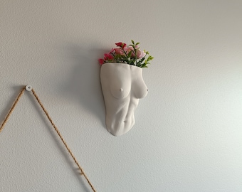 Könnte beinhalten: Weißer Keramik-Pflanztopf in Form eines Torsos mit rosa Blumen darin. Der Topf ist an einer weißen Wand befestigt.