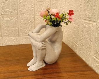 Könnte beinhalten: Eine weiße Keramikvase in Form einer sitzenden Person mit gekreuzten Beinen, die einen Strauß rosa und roter Blumen hält.