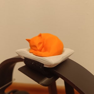 Puede incluir: Figura de gato naranja impresa en 3D durmiendo en una almohada blanca, unida a un soporte de auriculares negro.
