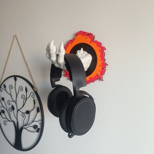 May include: A black gaming headset suspended from a wall-mounted headphone holder. The holder is a white skeletal hand emerging from a fiery orange and red circle. A black metal tree decoration hangs on the wall.