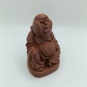 May include: A brown figurine of a seated Buddha with a cartoon character's face, wearing glasses and a bow tie. The figure is in a meditative pose, with a rounded belly and detailed robes. The base has a decorative pattern.