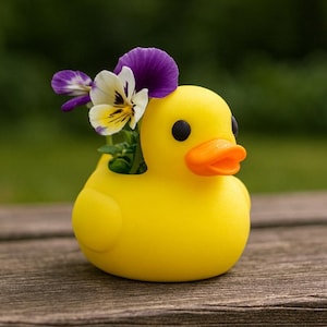 Op de afbeelding: Een gele eendvormige bloempot met een oranje snavel en zwarte ogen. De bloempot bevat paarse en witte viooltjes. De eend staat op een houten oppervlak, met een vage groene achtergrond.