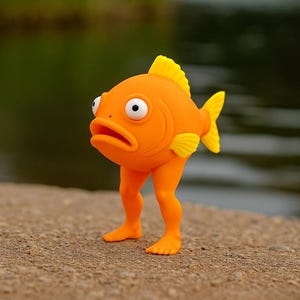 May include: An orange fish figurine with yellow fins and a yellow dorsal fin stands on two orange legs. The fish has large, surprised-looking eyes and an open mouth. The background is a blurred view of water and greenery.