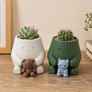 Puede incluir: Dos macetas de cerámica caprichosas, una blanca y otra verde, cada una con una pequeña suculenta y un cactus. La maceta blanca sostiene un osito de peluche marrón, y la maceta verde sostiene una figura de gato azul. Ambas macetas tienen caras sonrientes.