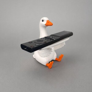 White 3D printed goose remote holder sitting on a gray studio background, holding a black TV remote across its wings with orange beak and feet.