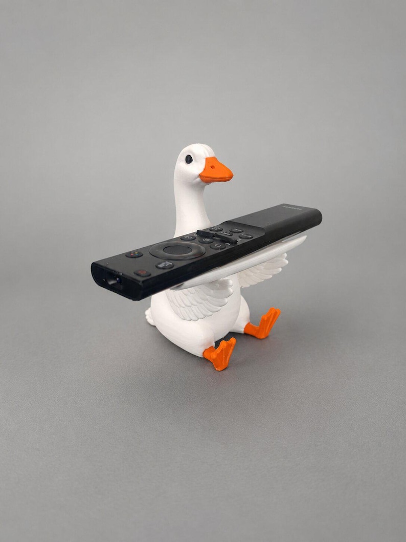 White 3D printed goose remote holder sitting on a gray studio background, holding a black TV remote across its wings with orange beak and feet.