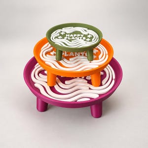 Peut inclure: Un empilement de trois supports pour plantes ronds de couleur vert olive, orange et magenta. Chaque support possède trois pieds courts et un motif ondulé blanc sur la surface. Le support supérieur vert olive affiche le texte blanc HAPPY PLANTS.