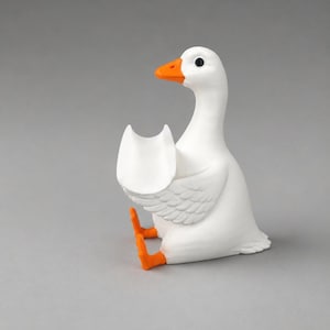 May include: A white goose figurine with a bright orange beak and orange feet. The goose is in a seated position with its wings slightly raised, forming a small, open, U-shaped cradle or holder in front of its chest. The surface is smooth and matte.
