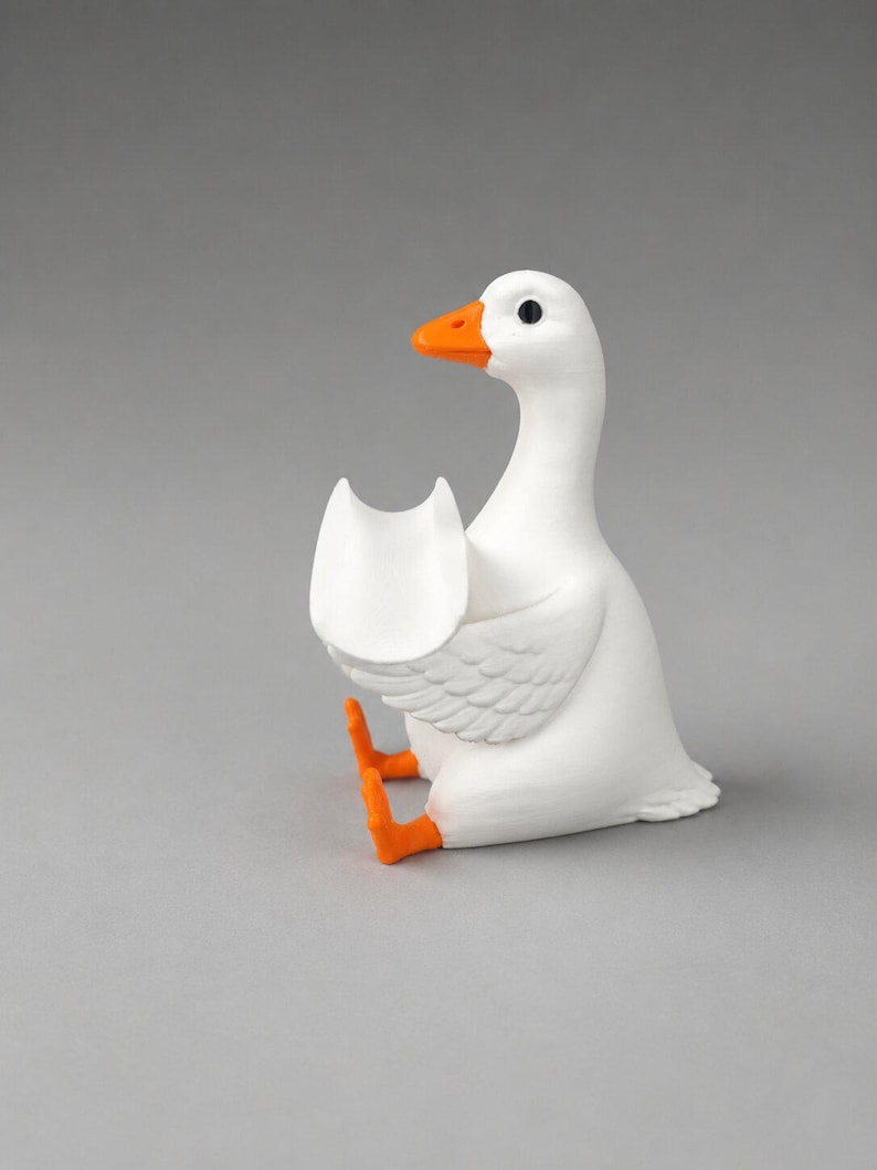 May include: A white goose figurine with a bright orange beak and orange feet. The goose is in a seated position with its wings slightly raised, forming a small, open, U-shaped cradle or holder in front of its chest. The surface is smooth and matte.