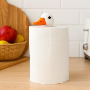 3D printed duck paper towel holder with magnetic head, shown on a kitchen counter with a basket of apples and a toaster in the background.