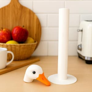 3D printed goose paper towel holder base with removable magnetic goose head, displayed on a kitchen counter with a cutting board, fruit basket, and toaster in the background.