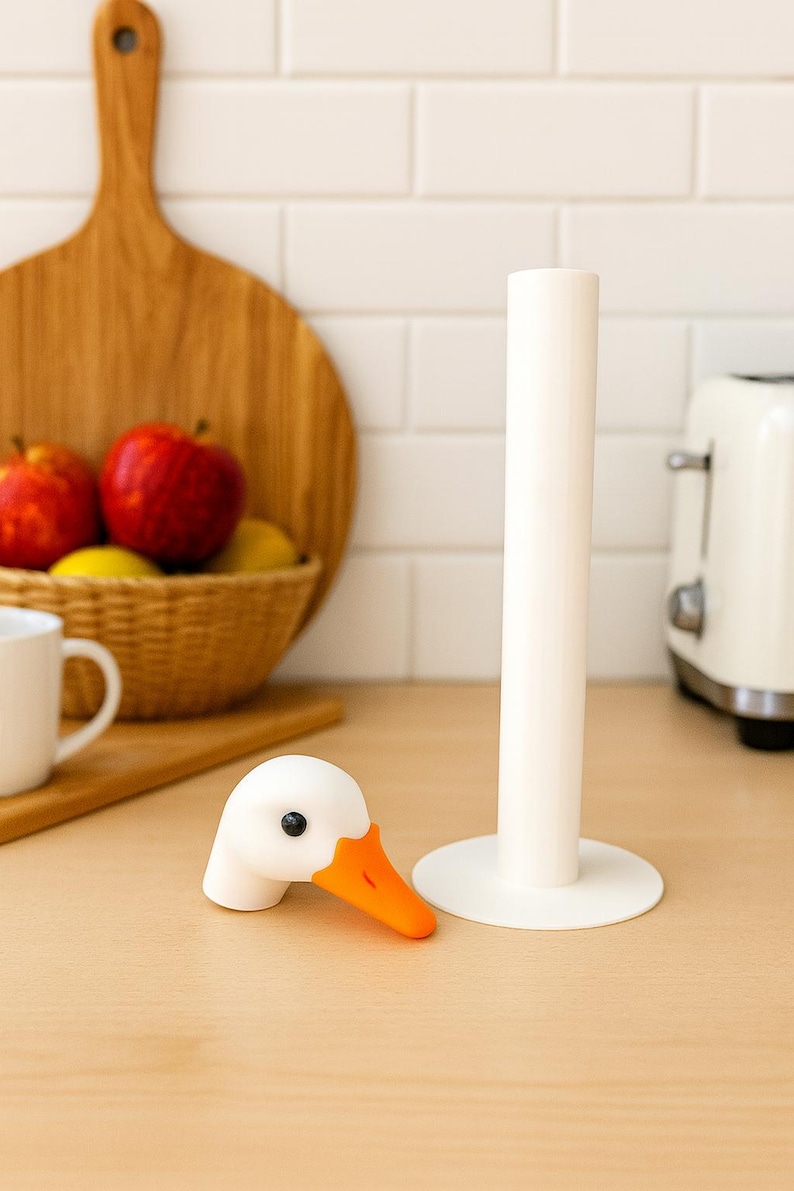 3D printed goose paper towel holder base with removable magnetic goose head, displayed on a kitchen counter with a cutting board, fruit basket, and toaster in the background.
