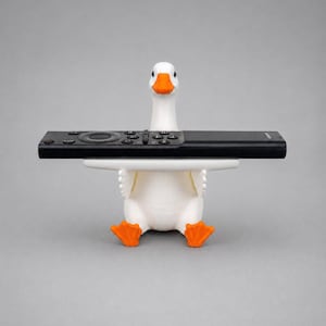 White 3D printed goose remote holder sitting on a gray studio background, holding a black TV remote across its wings with orange beak and feet.