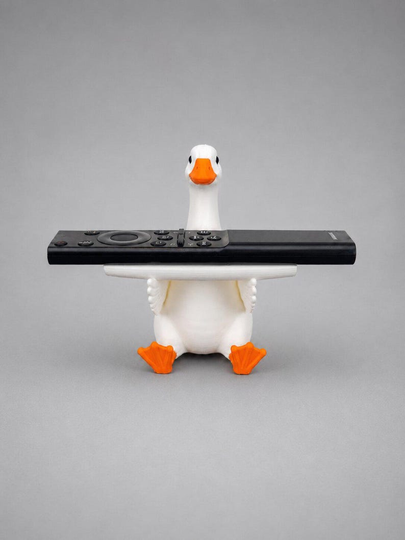 White 3D printed goose remote holder sitting on a gray studio background, holding a black TV remote across its wings with orange beak and feet.