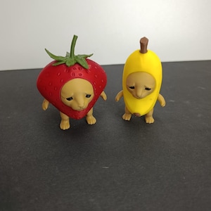 Pode incluir: Duas pequenas figuras de criaturas bege com expressões tristes. Uma criatura veste um traje de morango vermelho com um caule verde, e a outra veste um traje de banana amarela com um caule castanho. Ambas as figuras têm braços e pernas.