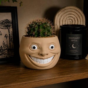 Op de afbeelding: Een grillige, beige gezichtsvormige plantenpot met een cactus. De pot heeft grote, witte ogen en een brede, grijnzende mond. Een zwarte kaars met de tekst "Midnight Spell" en een skeletdinosaurus staan ook op de plank.