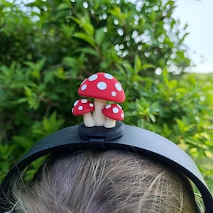 Può includere: Una fascia nera con un gruppo di tre figurine di funghi rossi e bianchi. I funghi hanno pois bianchi sui loro cappelli rossi. Lo sfondo è un fogliame verde sfocato e un cielo blu.