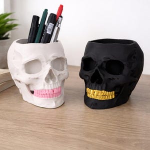 Può includere: Due portaoggetti a forma di teschio. Uno è bianco con denti rosa, che contiene penne e pennarelli. L'altro è nero con denti dorati. Entrambi sono realizzati in materiale duro e poggiano su una superficie di legno.