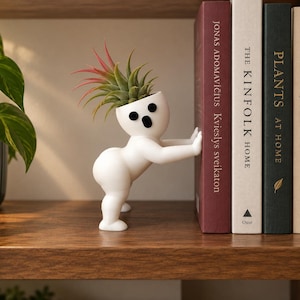 Peut inclure: Un pot de fleurs blanc en forme de personnage avec une expression surprise, doté de deux yeux noirs et d'une bouche ouverte. La figurine est positionnée pour soutenir une rangée de livres, avec une petite plante aérienne verte et rouge.