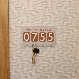 Könnte beinhalten: Ein rechteckiger Schlüsselhalter mit dem Text "KM Run This Year" und der Zahl "0755" in Braun. Der Schlüsselhalter hat vier Haken und einen Satz Schlüssel, der an einem Haken hängt. Der Schlüsselhalter ist an einer strukturierten, hellfarbigen Wand befestigt.