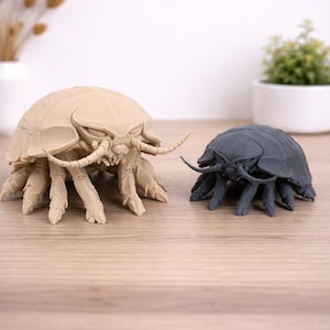May include: Two detailed insect sculptures. One is a light beige colour, and the other is a dark grey. Both have multiple legs, antennae, and a segmented body. They are placed on a wooden surface, with a white background.