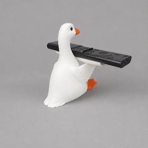 White 3D printed goose remote holder sitting on a gray studio background, holding a black TV remote across its wings with orange beak and feet.