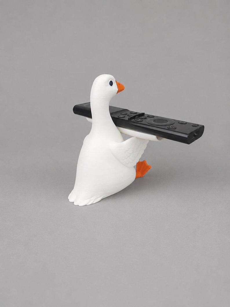White 3D printed goose remote holder sitting on a gray studio background, holding a black TV remote across its wings with orange beak and feet.