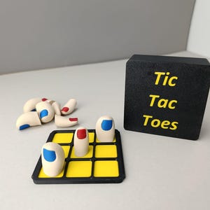 Może przedstawiać: Gra Tic Tac Toe z czarną planszą i żółtymi polami. Na planszy znajdują się białe, przypominające palce pionki z czerwonym i niebieskim lakierem do paznokci.