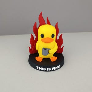 Könnte beinhalten: Gelbe Entenfigur mit orangefarbenem Schnabel und Füßen, die einen grauen Becher hält. Die Ente ist von roten Flammen umgeben und sitzt auf einem schwarzen Sockel mit dem weißen Text "THIS IS FINE".