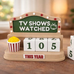 Puede incluir: Un contador de programas de televisión con las palabras "TV SHOWS WATCHED" sobre un fondo verde. El contador tiene cubos numéricos blancos para rastrear el número de programas vistos. Un pequeño recipiente de palomitas de maíz está a la izquierda. El texto "THIS YEAR" está en la base.