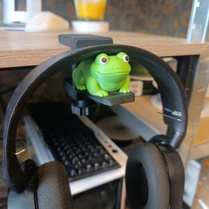 Könnte beinhalten: Ein grüner, froschförmiger Kopfhörerhalter ist an einem Schreibtisch befestigt, an dem ein schwarzer Kopfhörer hängt. Der Frosch hat große, runde Augen und befindet sich unter der Schreibtischkante. Im Hintergrund sind eine Computertastatur und ein Glas mit orangener Flüssigkeit zu sehen.