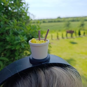 May include: A miniature cup of yellow noodles with two brown chopsticks sits atop a black headband. The small food item is in a white cup. The background is a blurred green field and blue sky.
