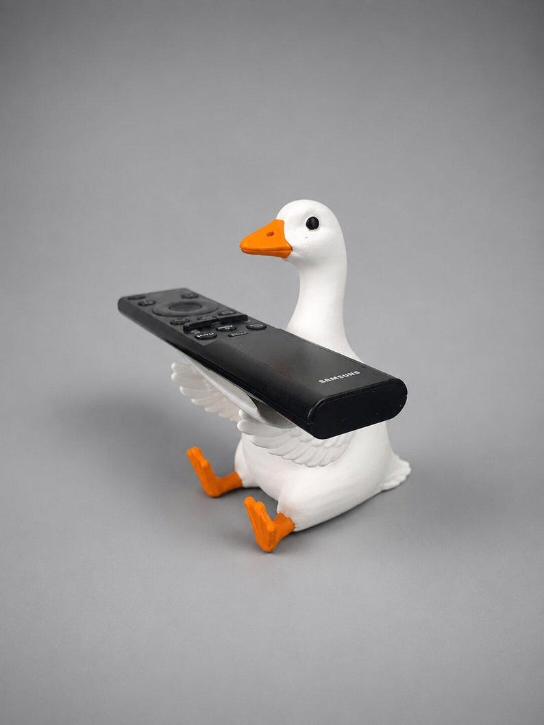 White 3D printed goose remote holder sitting on a gray studio background, holding a black TV remote across its wings with orange beak and feet.